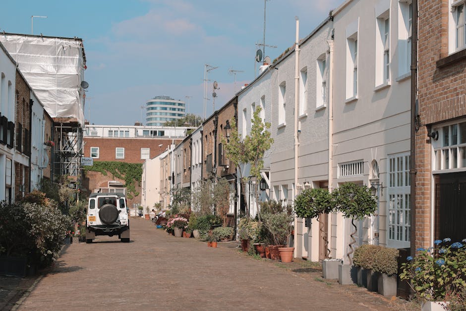 A narrow cobblestone street in Kilburn, lined with modern and traditional terraced houses featuring white and brick facades. The gardens in front of the homes are well-maintained, with a variety of po